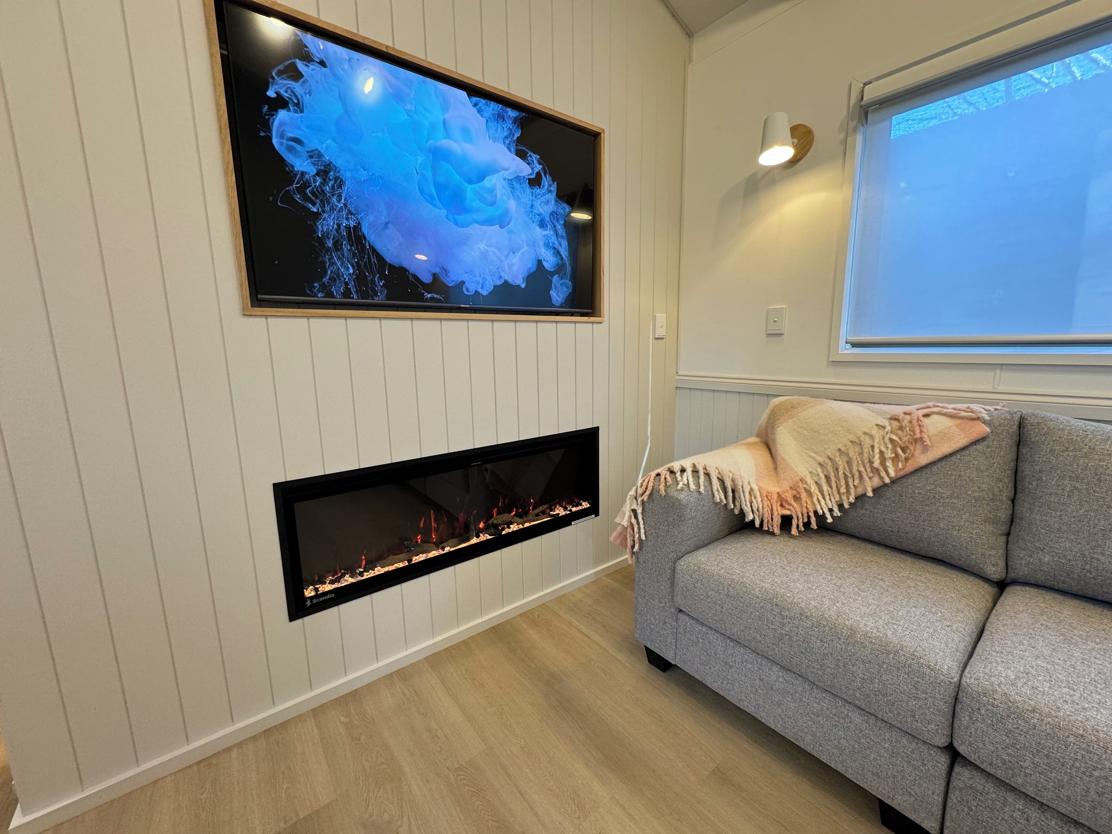 Una sala moderna con una televisión de pantalla plana sobre la chimenea, un sofá gris con una manta y una ventana con persianas.