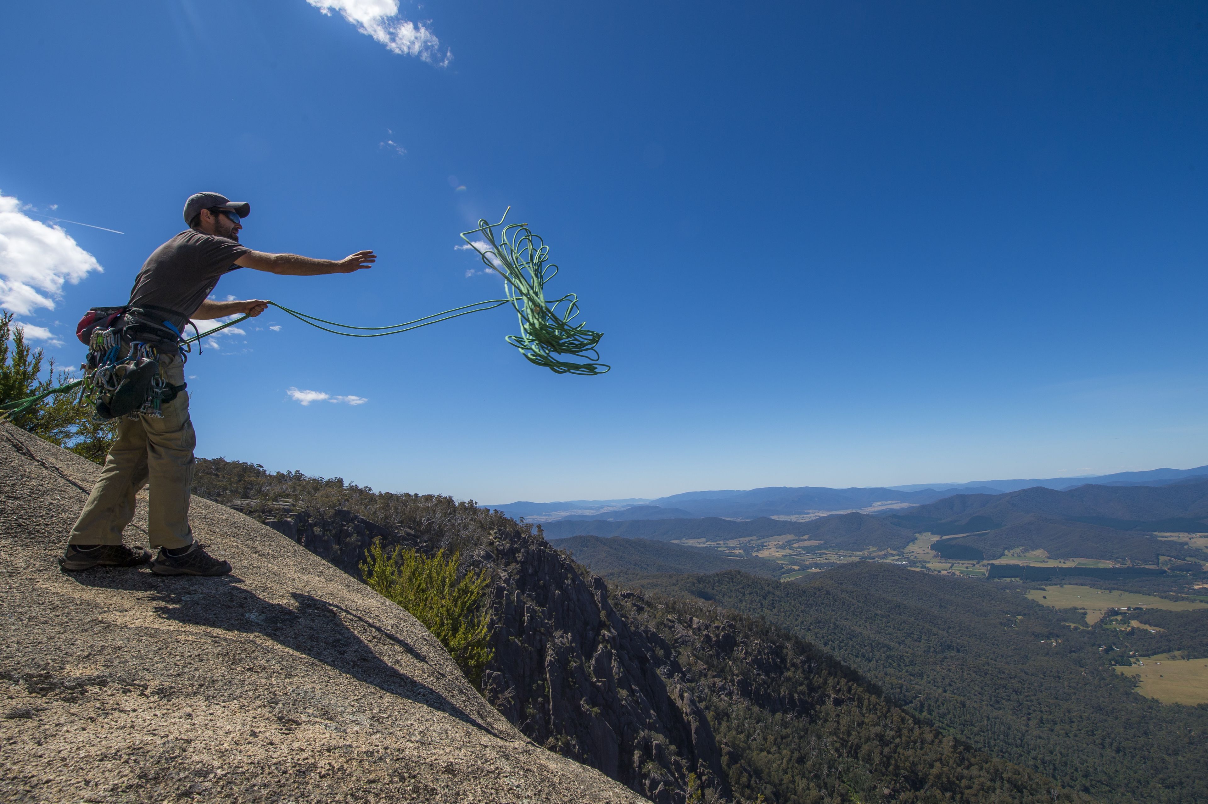 A person standing on a rocky outcrop with a rope, overlooking a valley with mountains and forests.