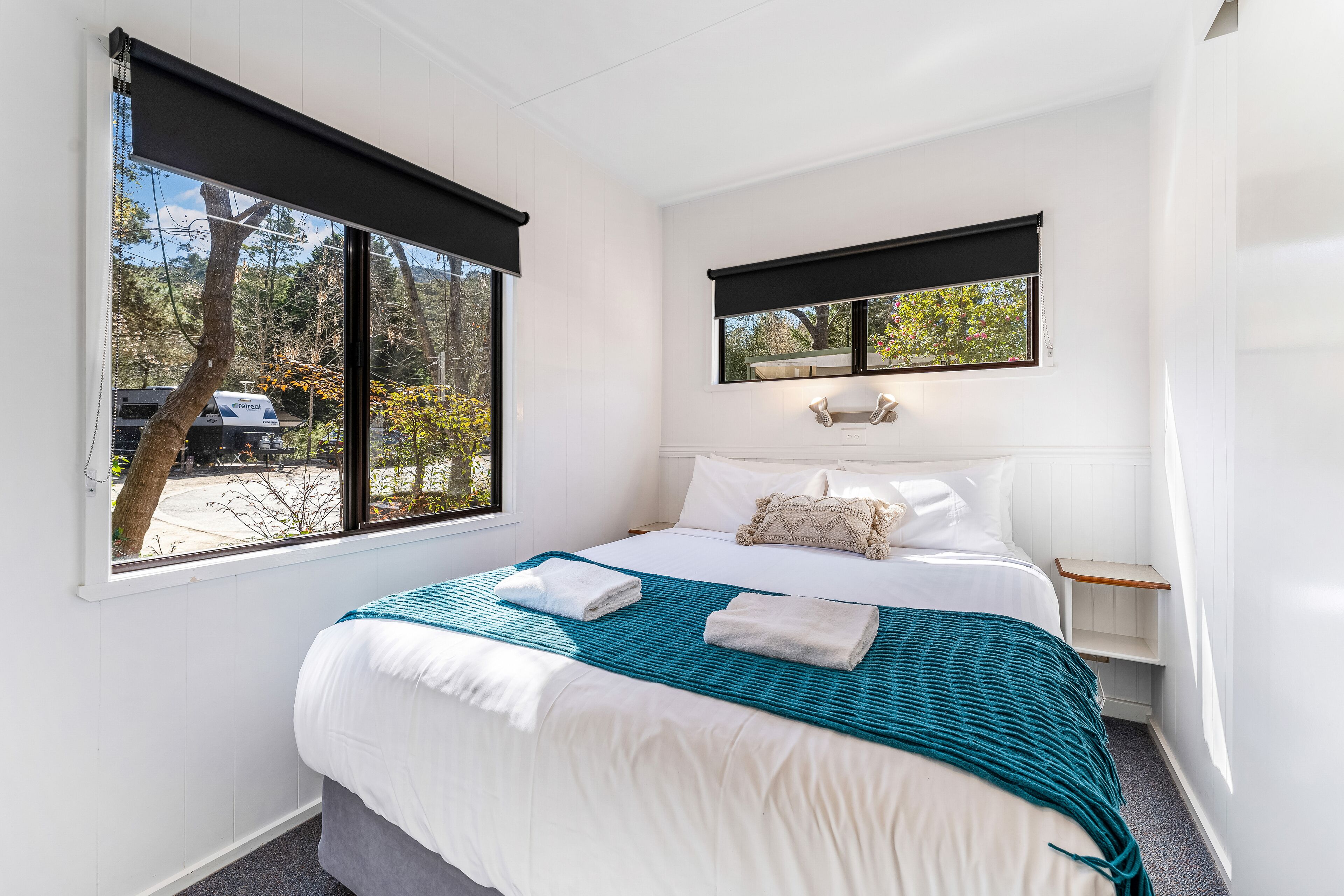 A bedroom with a bed, a window with a view of trees and a parked car, a bedside table with a lamp, and a small shelf.