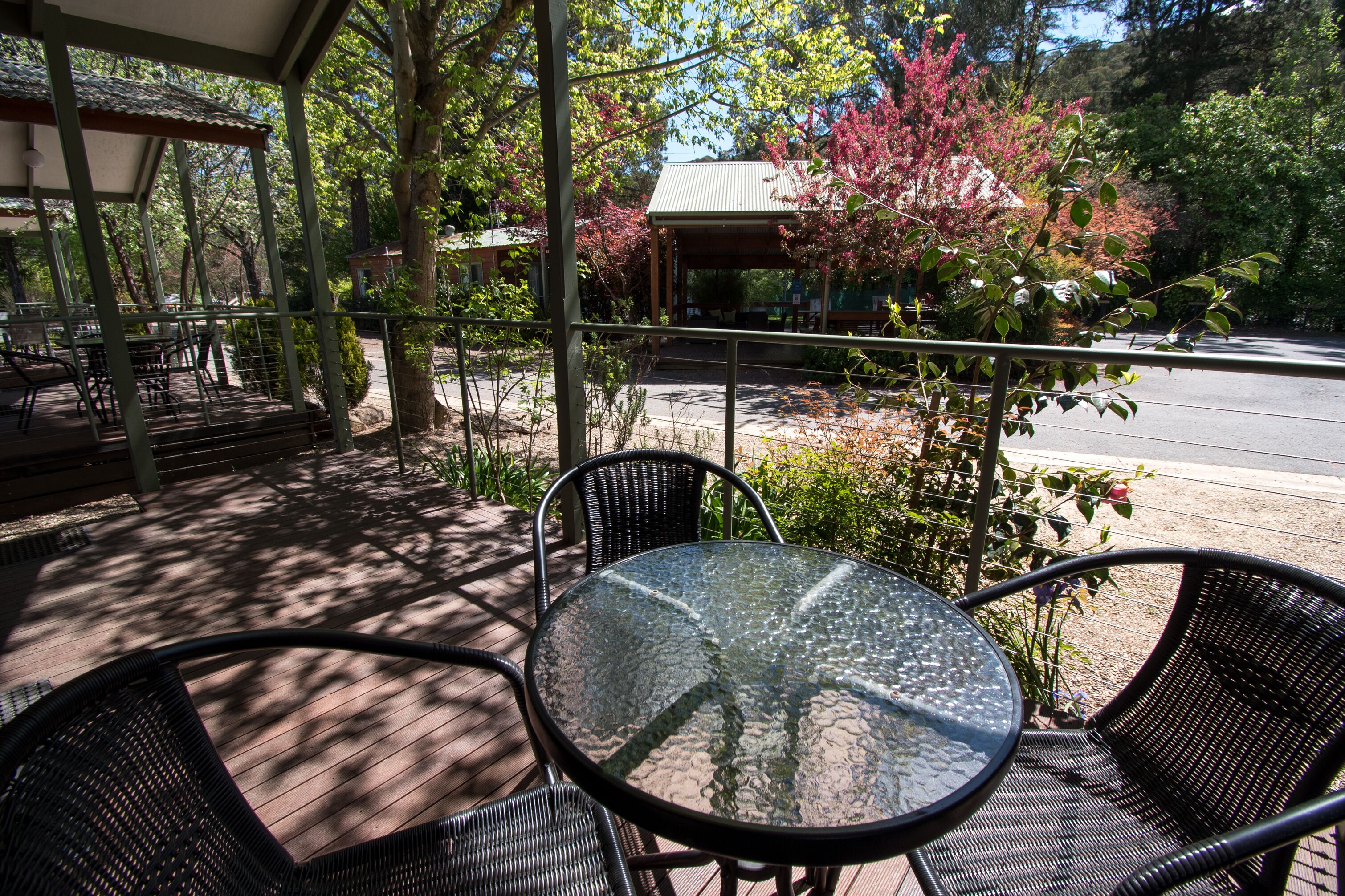 An outdoor patio with a round glass table and wicker chairs, surrounded by trees and a building with a green roof.