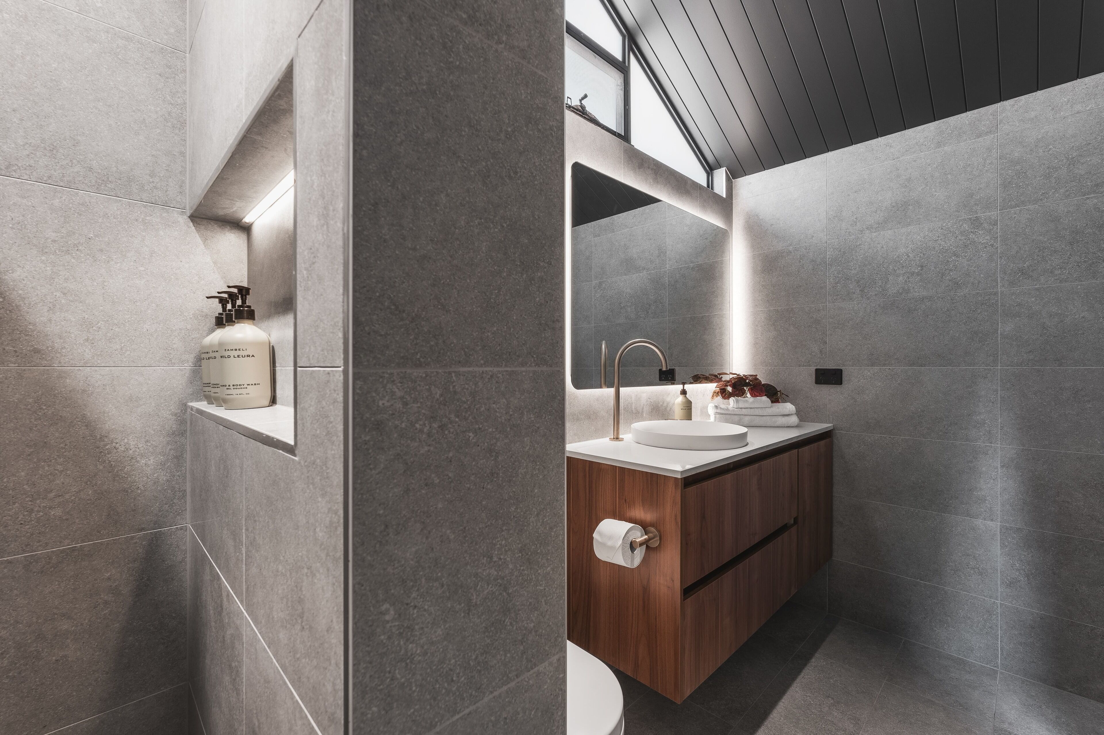 A modern bathroom with grey tiles, a wooden vanity, a white sink, and a large mirror.