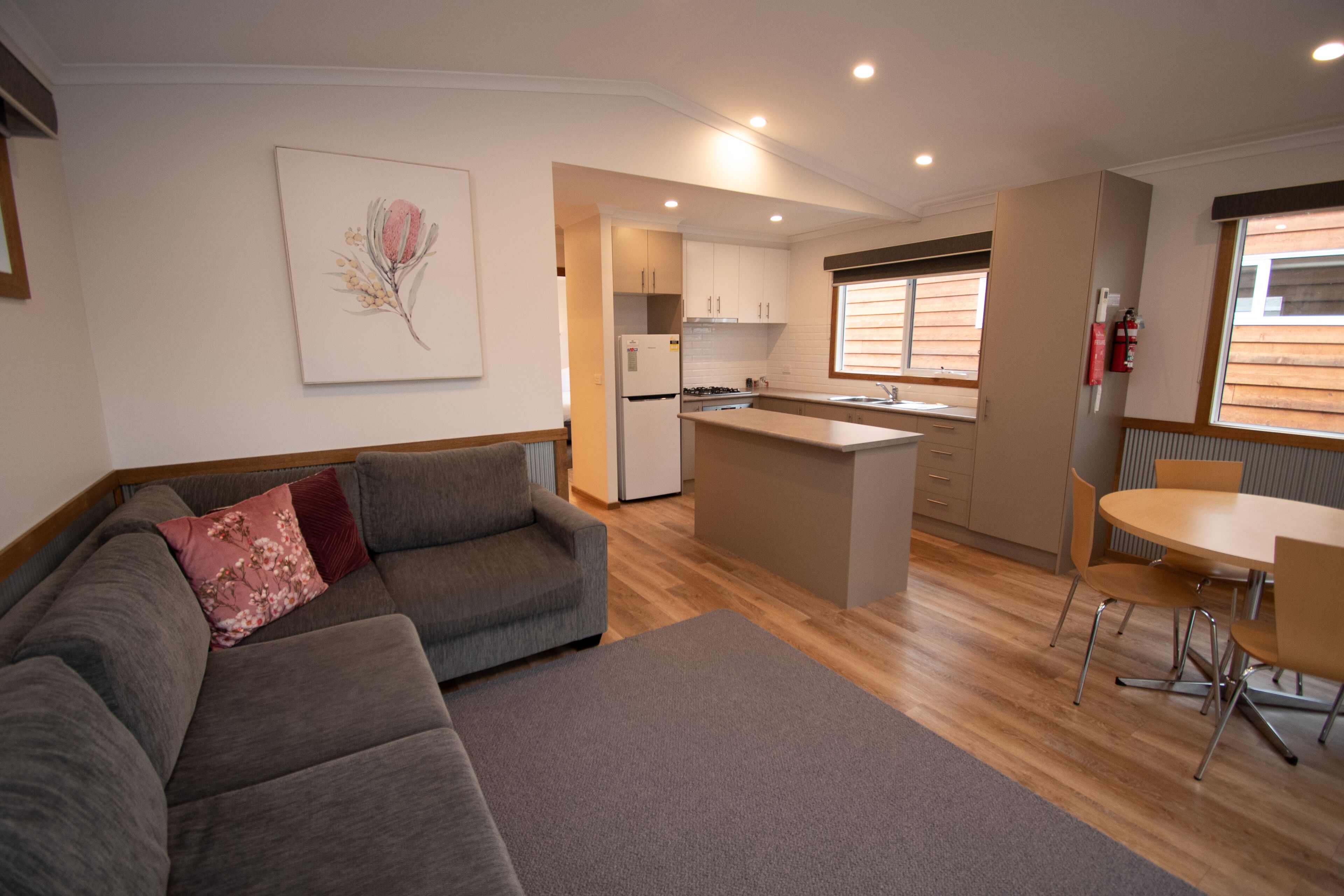 Una sala de estar moderna con un sofá gris, una pequeña mesa de comedor y una zona de cocina con armarios de madera y una isla integrada.