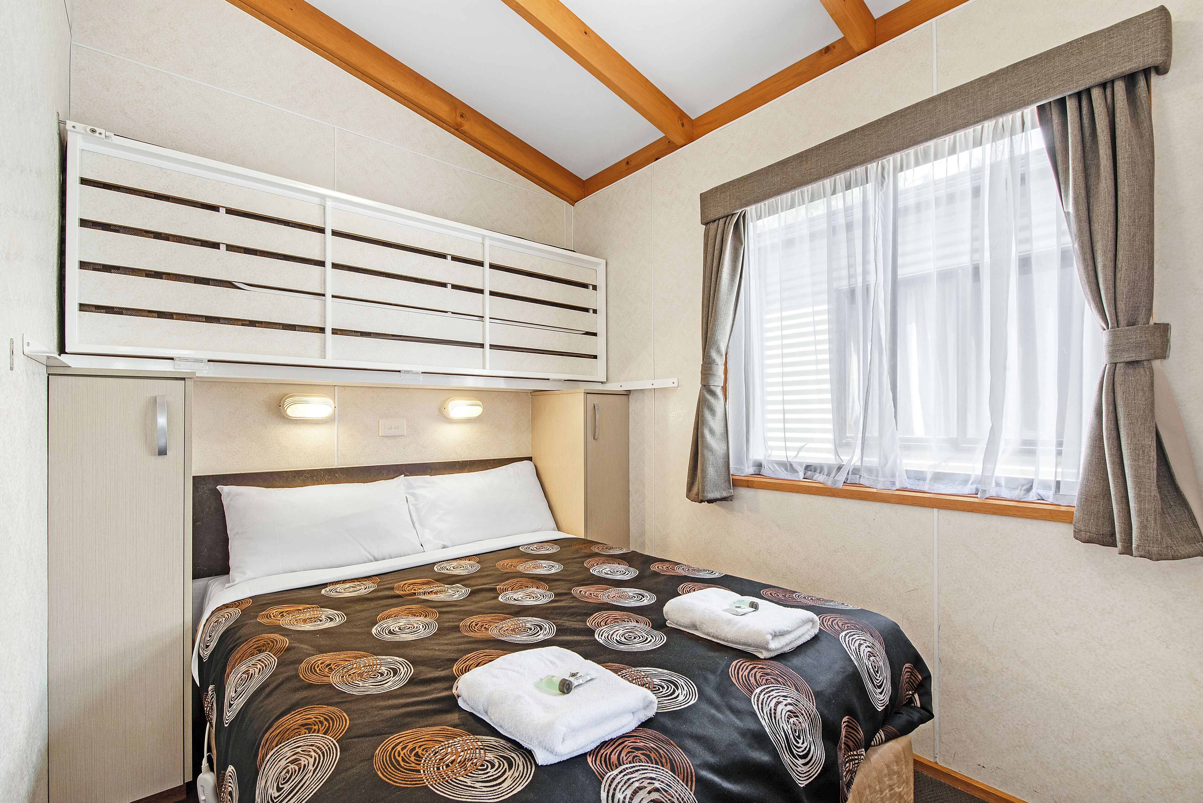 A single bed with a patterned bedspread, a headboard, and a window with curtains.