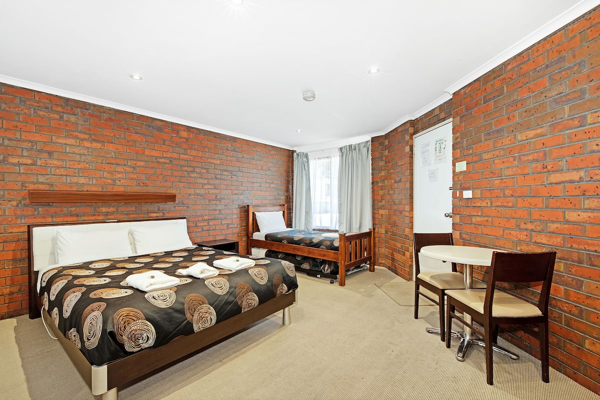 A hotel room with a brick wall, a bed with a patterned bedspread, a desk with a chair, and a whiteboard on the wall.