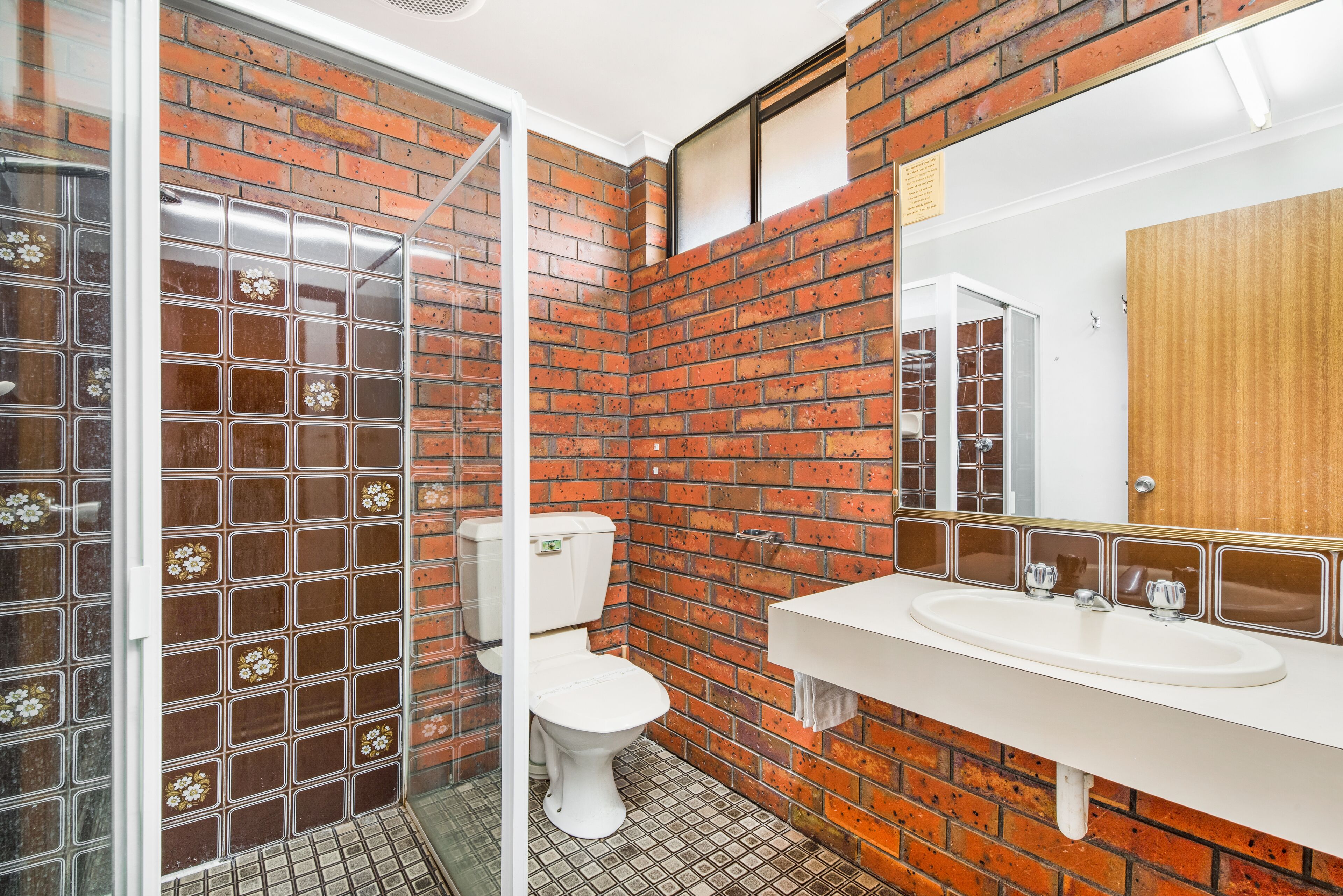 A bathroom with a brick wall, a glass shower enclosure, a white toilet, and a white sink with a mirror above it.