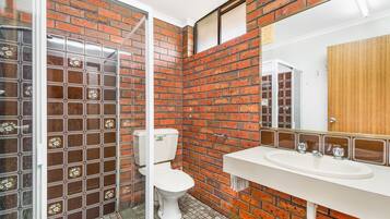 A bathroom with a brick wall, a glass shower enclosure, a white toilet, and a white sink with a mirror above it.