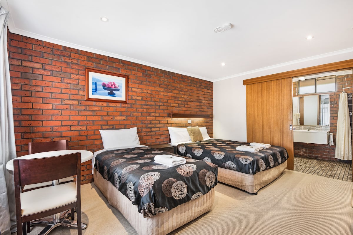 A hotel room with two beds, a wooden headboard, a brick accent wall, a framed picture, a small table, and a chair.