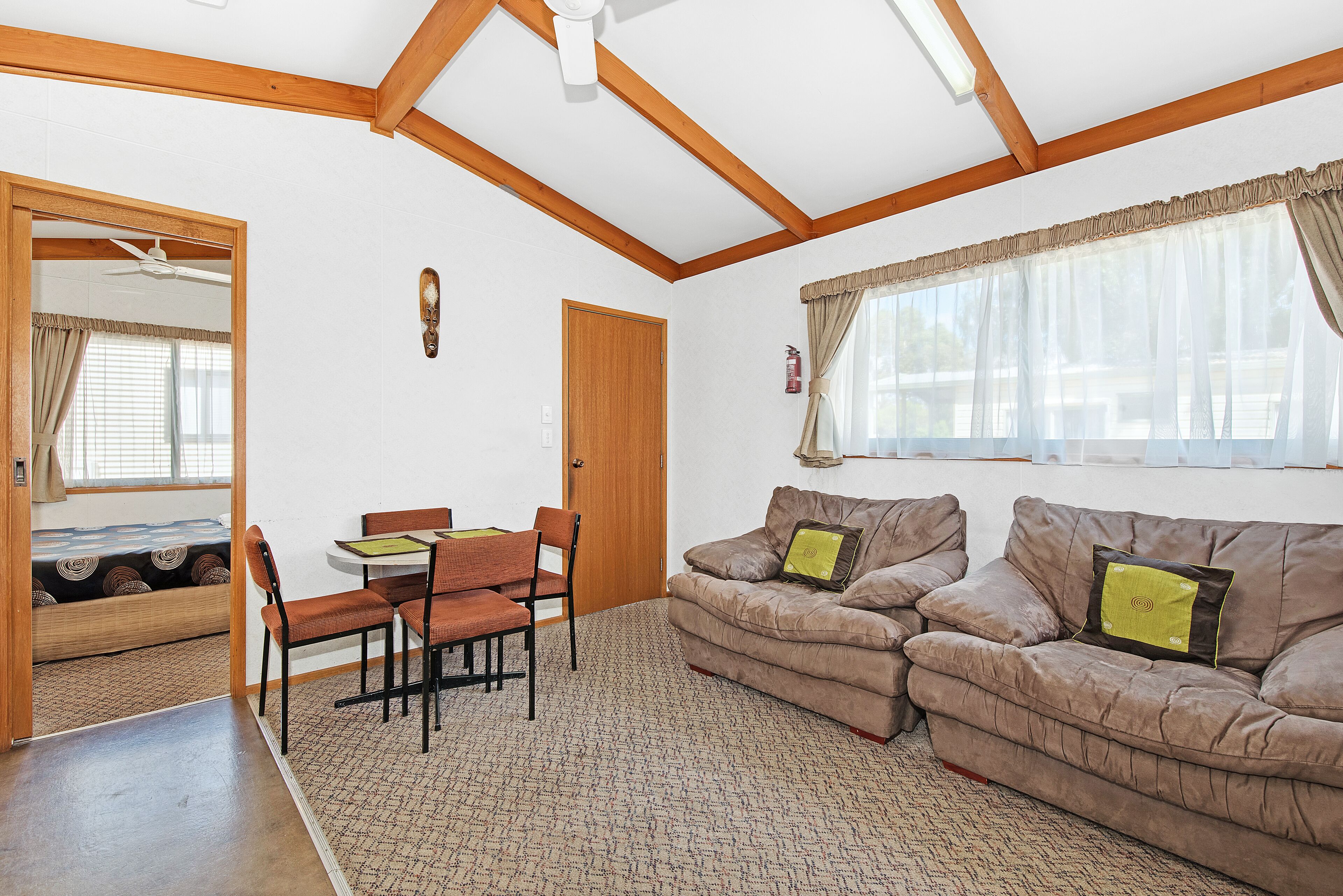 A living room with a sofa set, a dining table with chairs, a wooden ceiling, and a window with curtains.