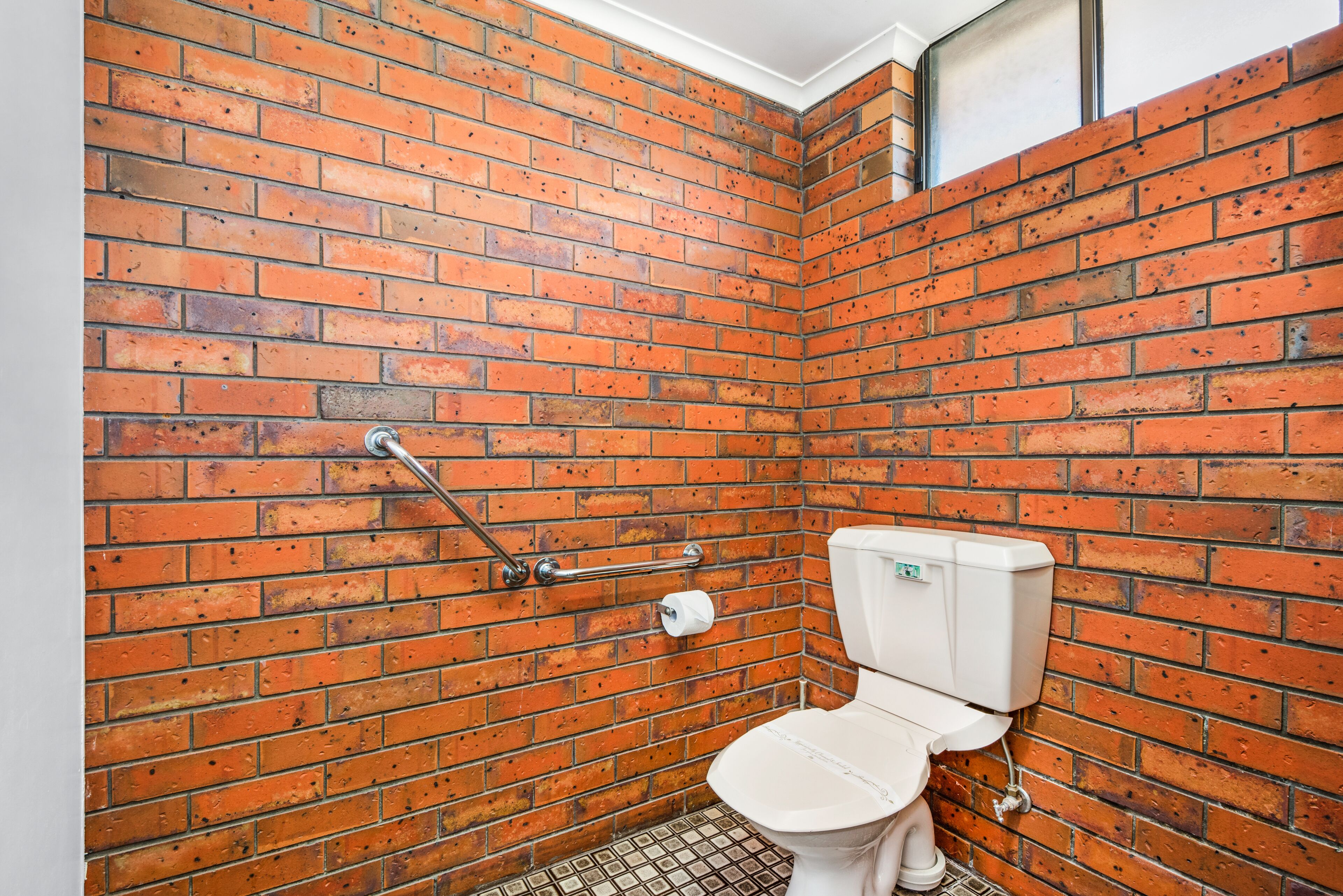 Ein kleines, abgeschlossenes Badezimmer mit Toilette, einer Haltestange und einer roten Backsteinwand.