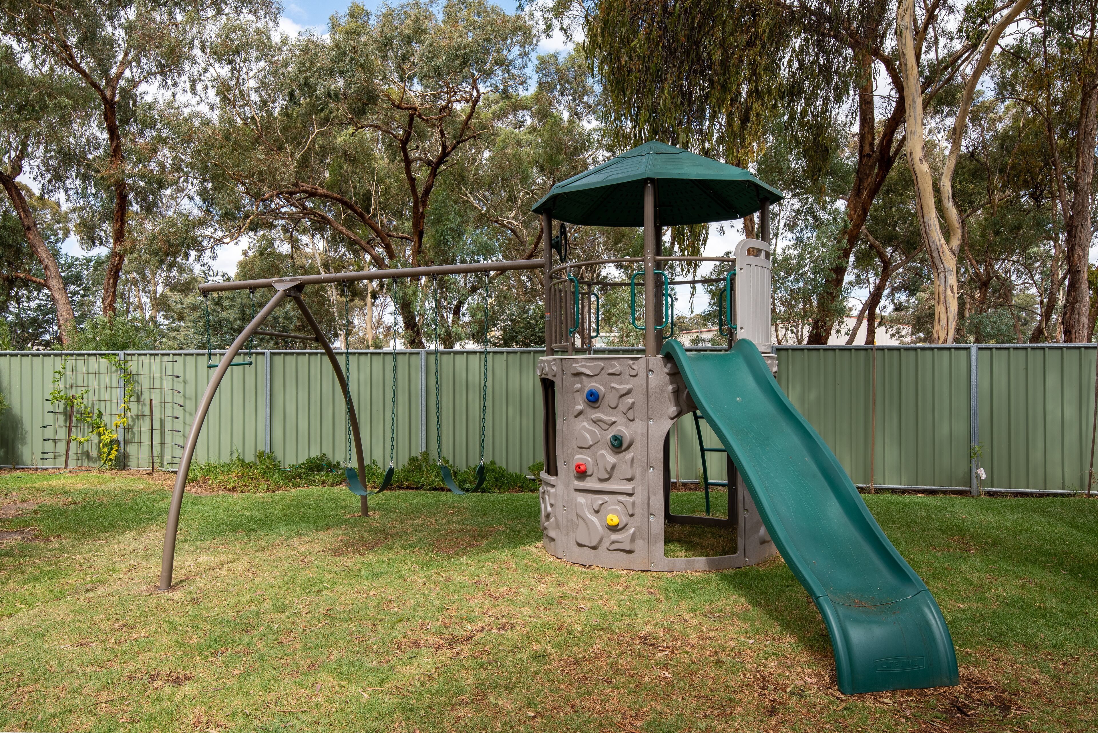A playground with a slide and climbing wall, surrounded by trees and a fence.