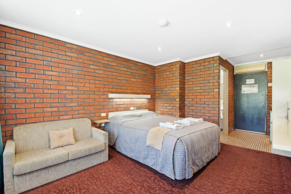 A hotel room with a brick wall, a bed with a blue bedspread, a beige sofa, and a red carpet.