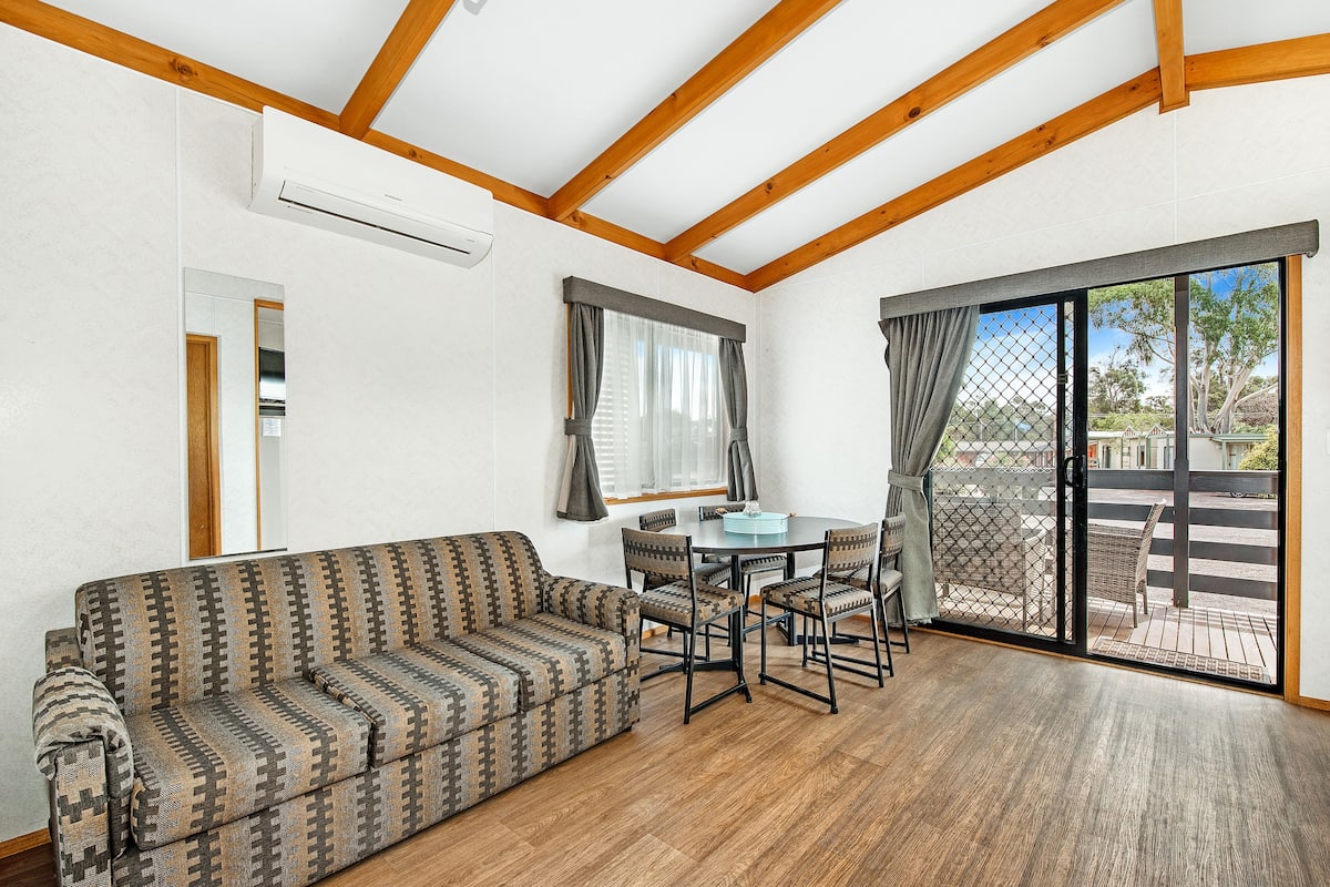 A room with a patterned sofa, a dining table with chairs, a sliding glass door leading to an outdoor area, and wooden beams on the ceiling.