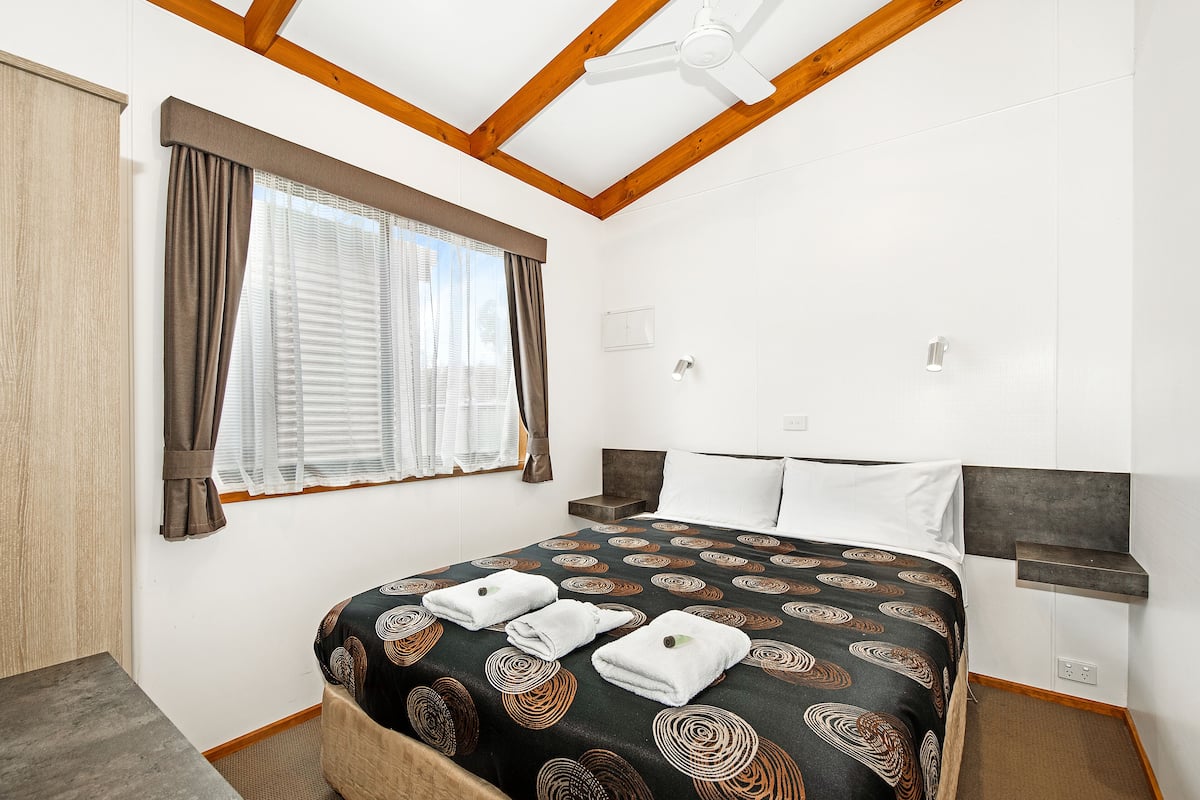 A neatly made bed with a patterned bedspread and three folded towels. There is a window with curtains, a ceiling fan, and a bedside table with a lamp.