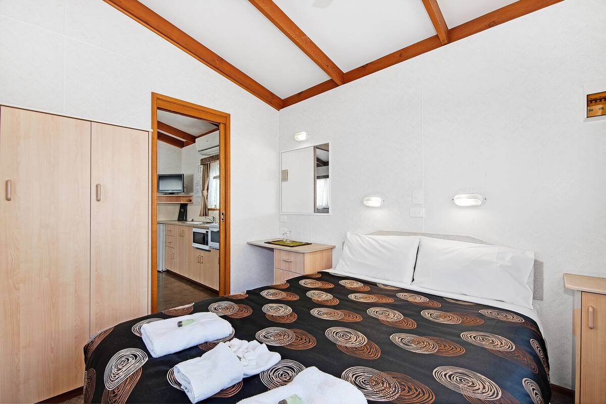 A hotel room with a bed, a wooden wardrobe, a small desk with a mirror, and a bathroom visible through an open door.