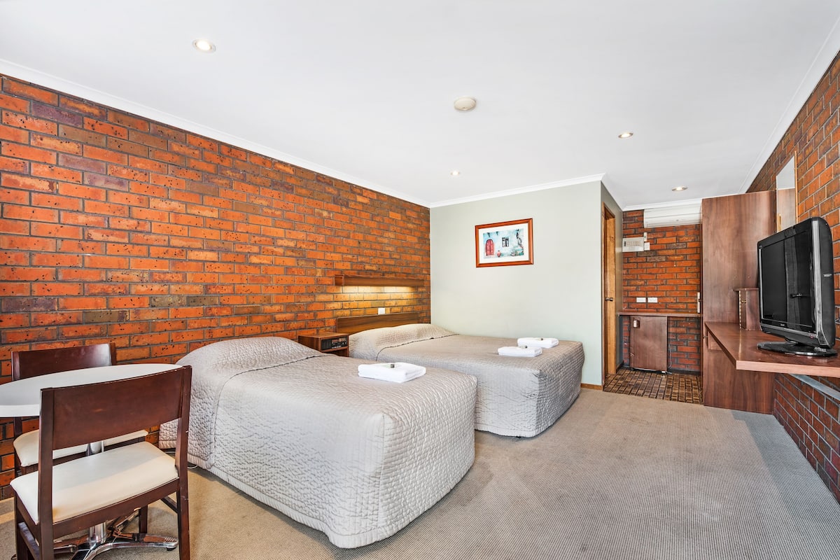 A hotel room with a brick wall, two beds, a TV, a desk, and a chair.