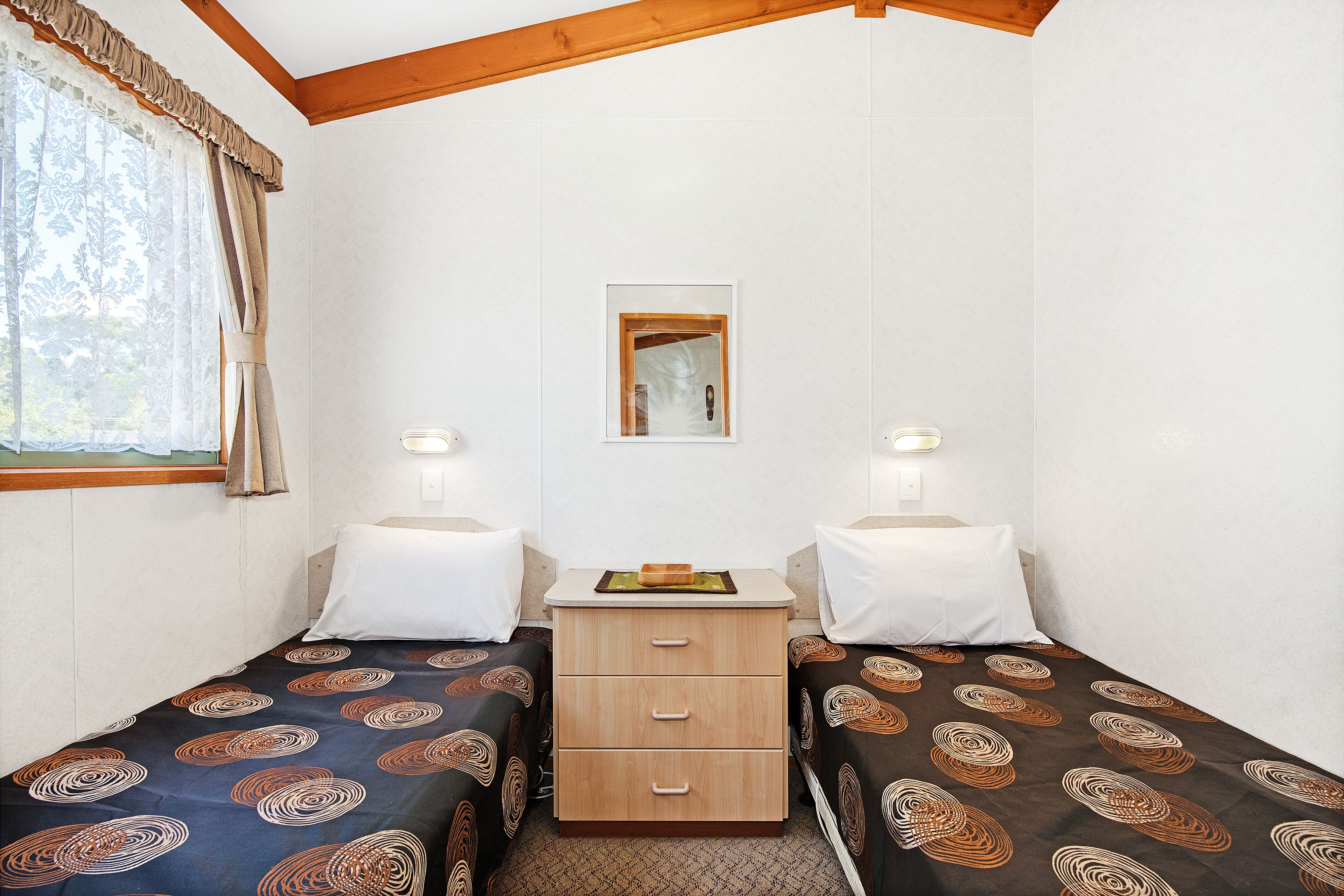 A small, clean room with two single beds, a wooden nightstand, a mirror, and two bedside lamps.