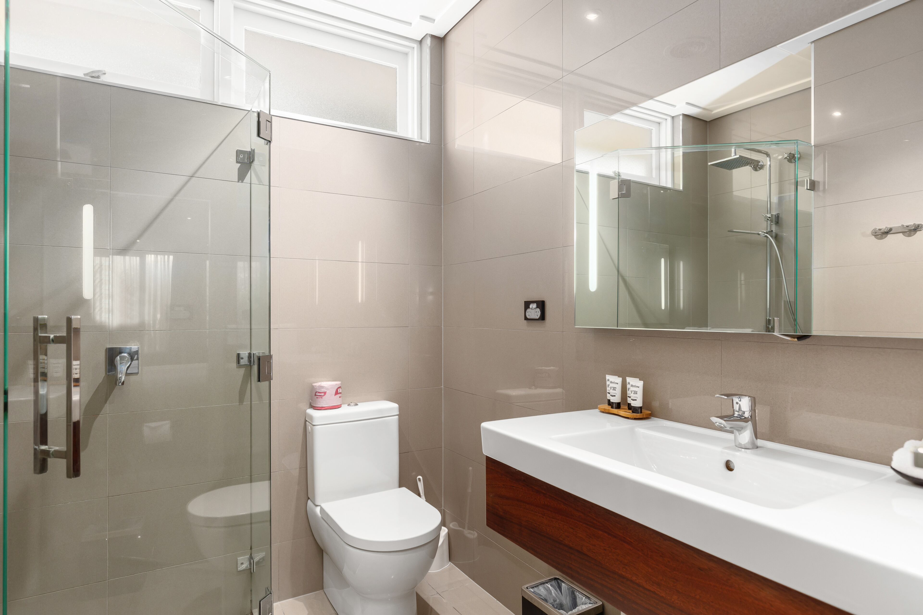 A modern bathroom with a glass-enclosed shower, a white toilet, and a large white sink with a wooden cabinet below.
