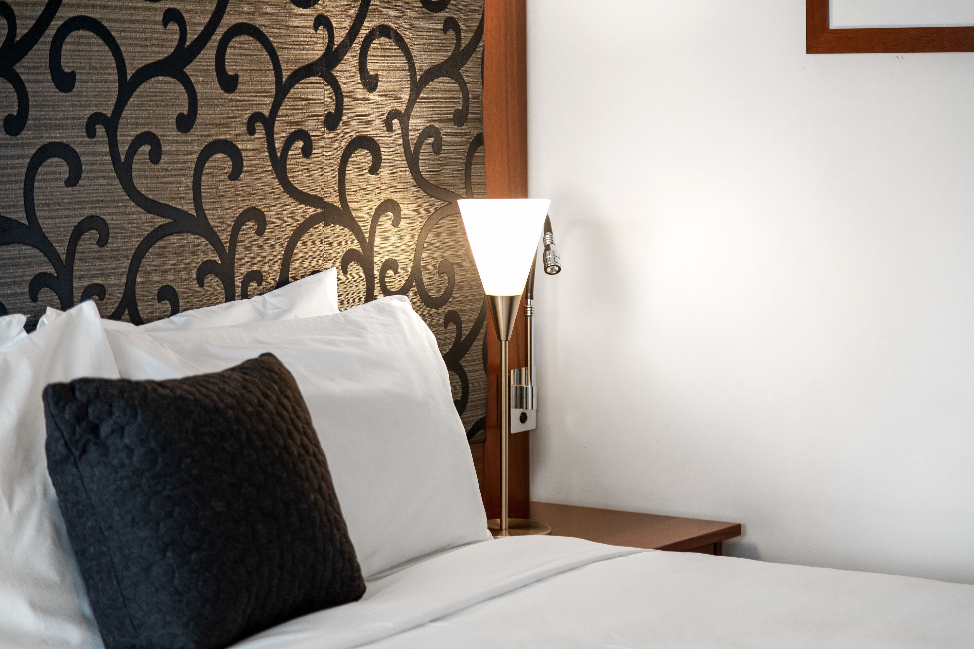 A neatly made bed with white and dark pillows, a bedside lamp, and a patterned wall.