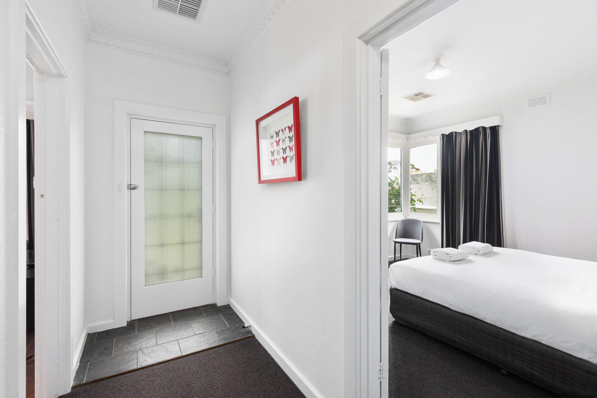 A hotel room with a bed, a chair, a window with curtains, a door with a frosted glass panel, and a framed picture on the wall.