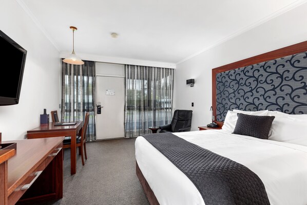 A hotel room with a large bed, a desk, a chair, a TV, and a view through the window. - The Commodore (Mount Gambier)