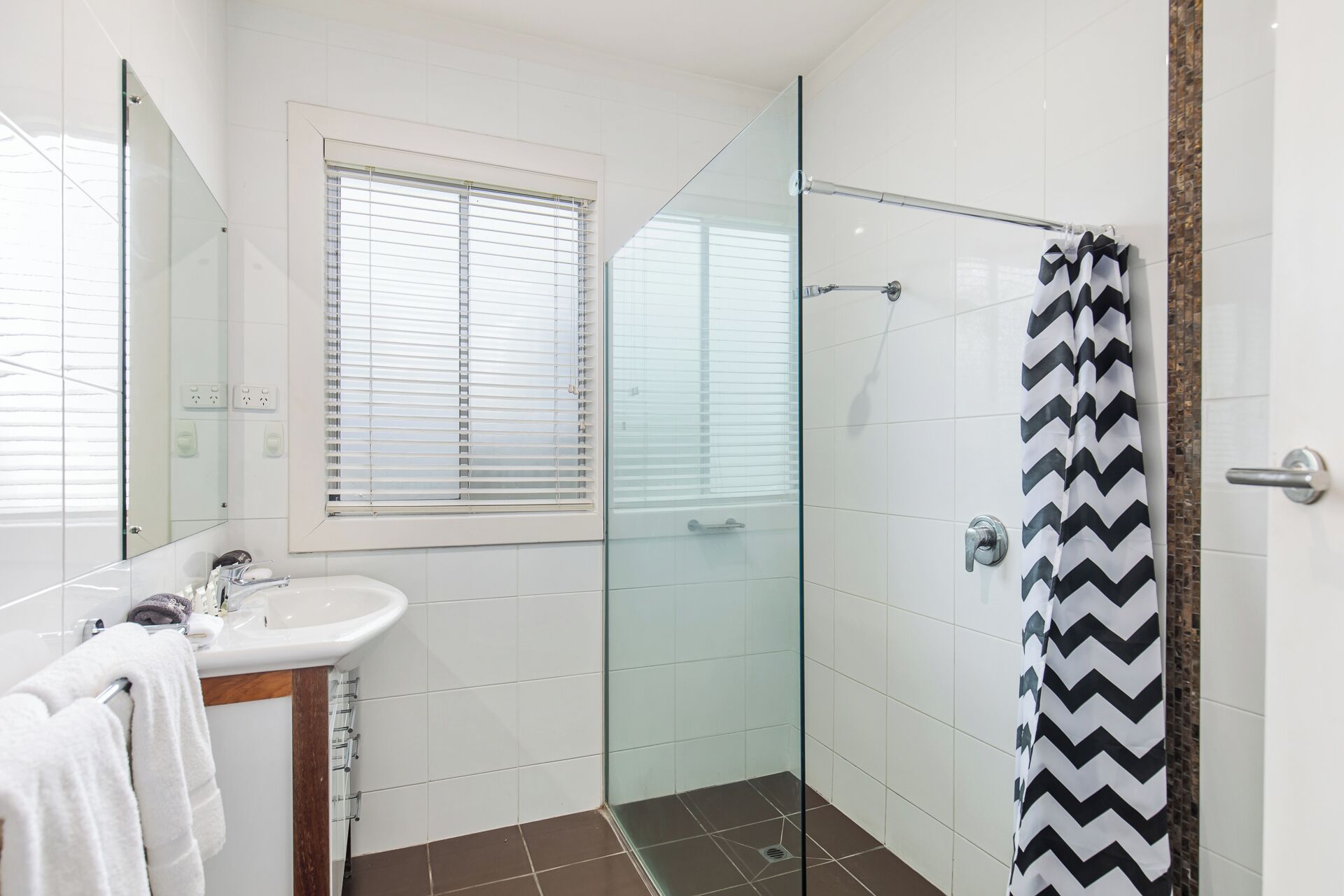 A bathroom with a glass shower enclosure, a sink with a mirror, and a window with blinds.