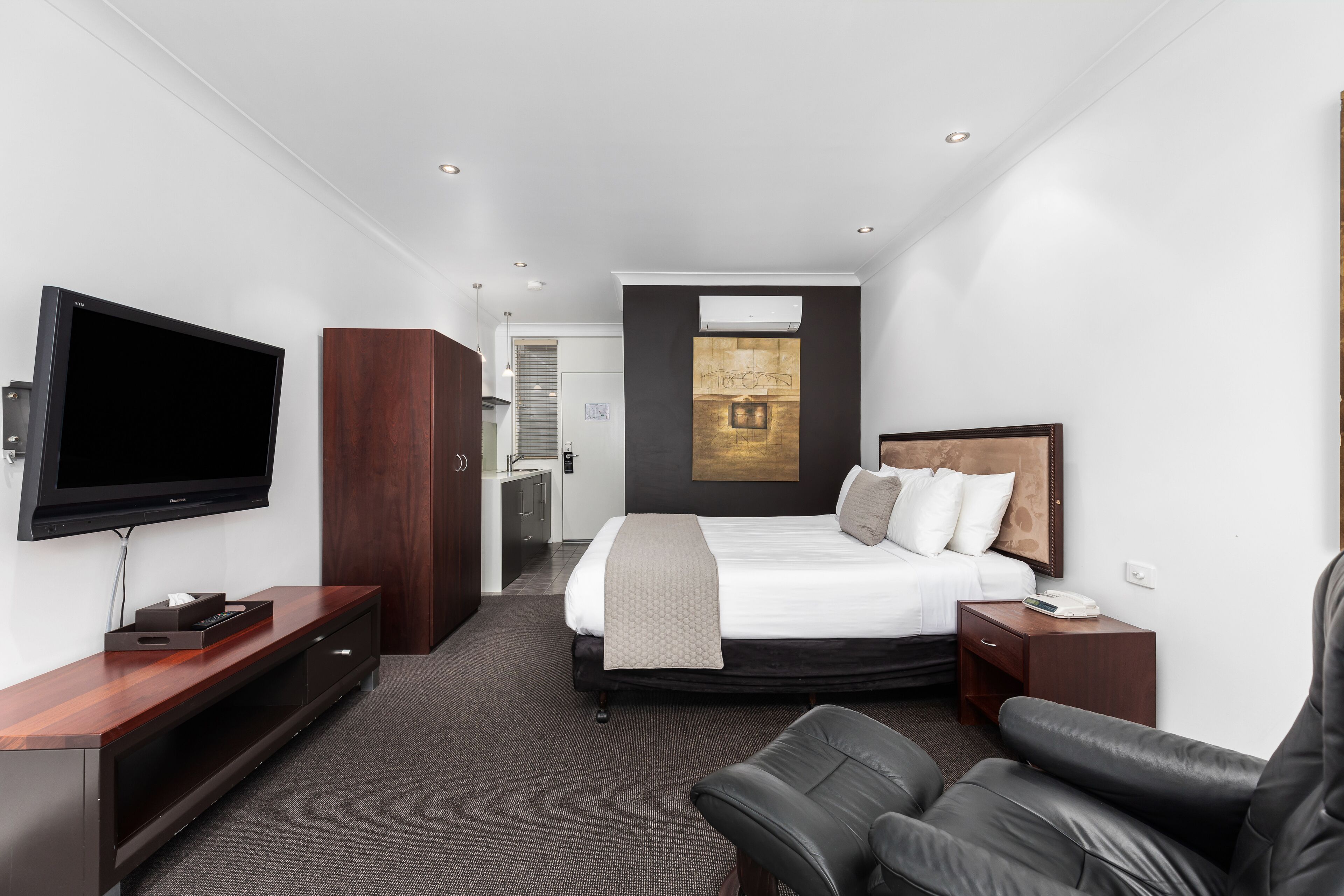 Une chambre d’hôtel moderne dotée d’un grand lit, d’un téléviseur à écran plat, d’un meuble de rangement en bois, d’un fauteuil en cuir et d’une petite table de chevet.