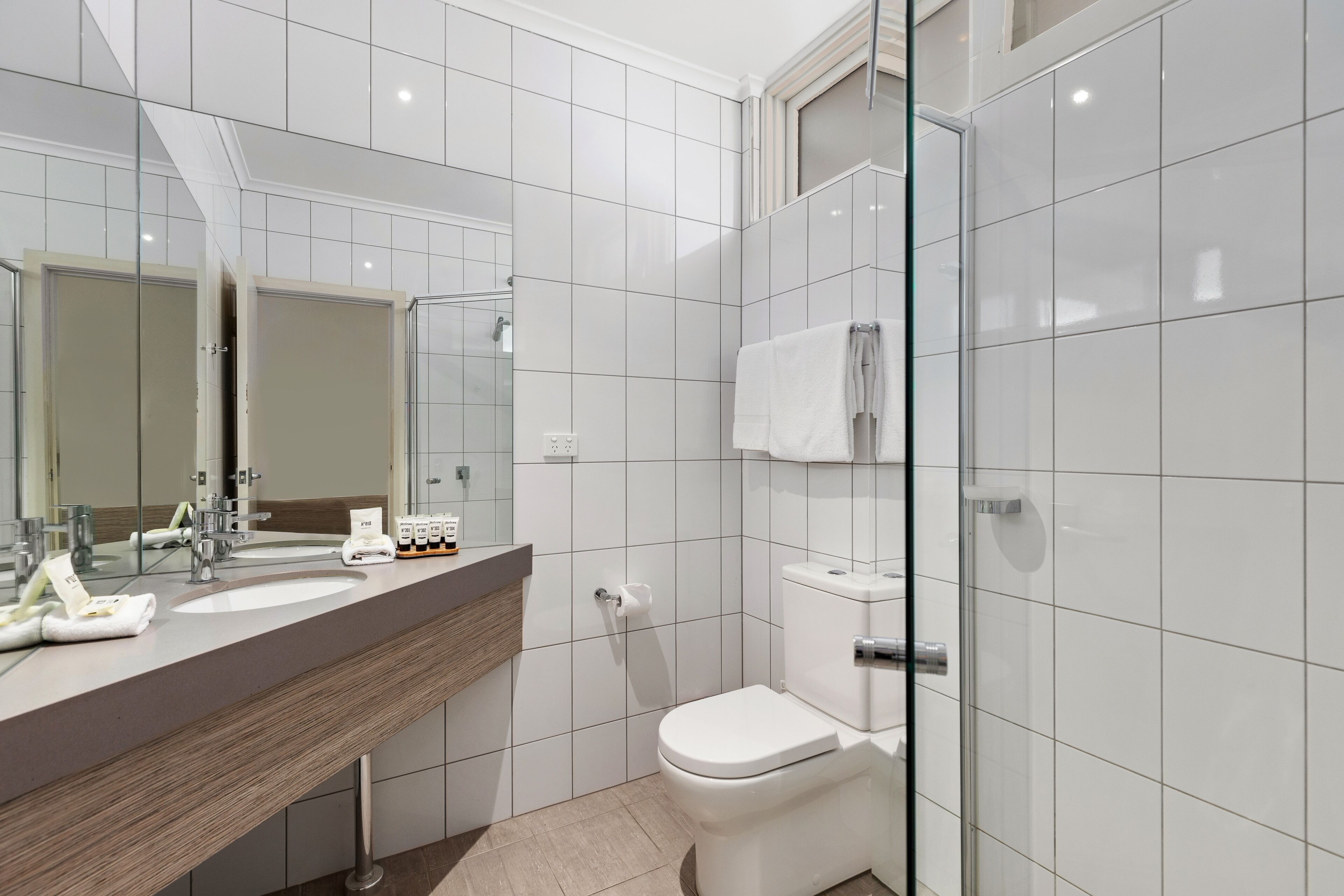 A modern bathroom with a large mirror, two sinks, a toilet, and a glass shower enclosure.