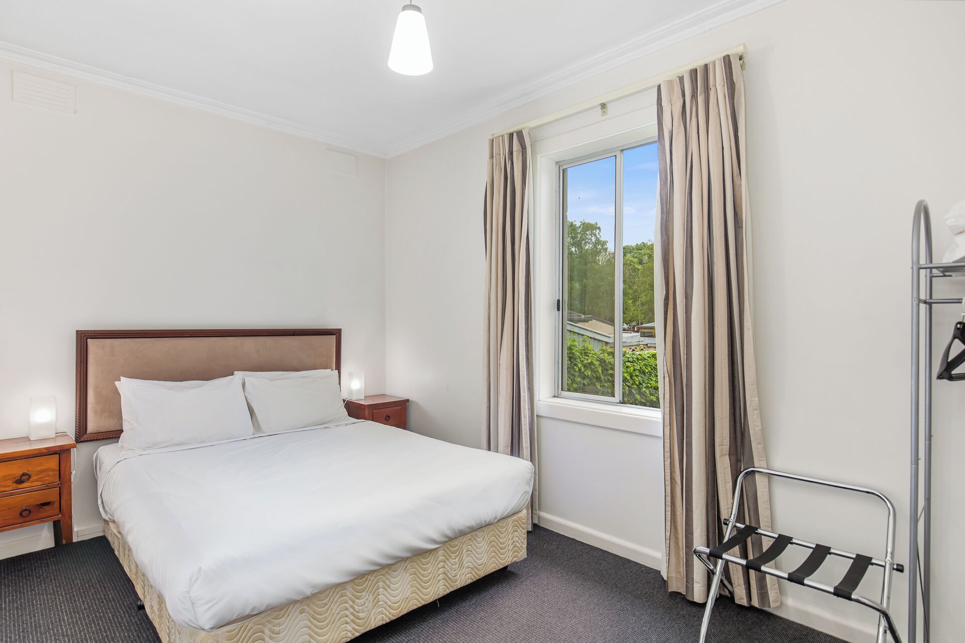A hotel room with a bed, a nightstand, a window with curtains, and a metal rack.