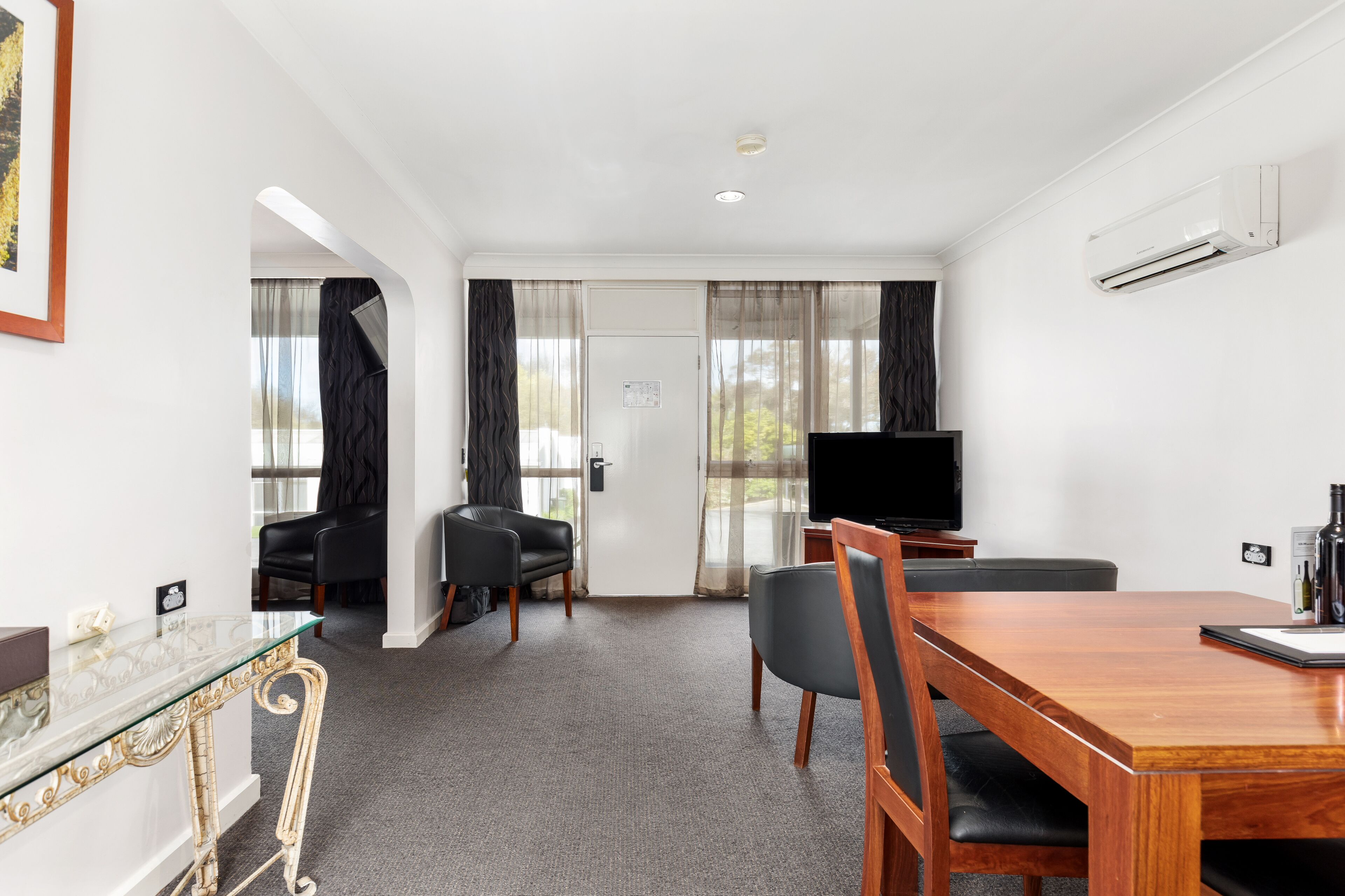 A hotel room with a wooden dining table, two black chairs, a glass coffee table, a flat-screen TV, and a large window with sheer curtains.