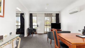 A hotel room with a wooden dining table, two black chairs, a glass coffee table, a flat-screen TV, and a large window with sheer curtains.