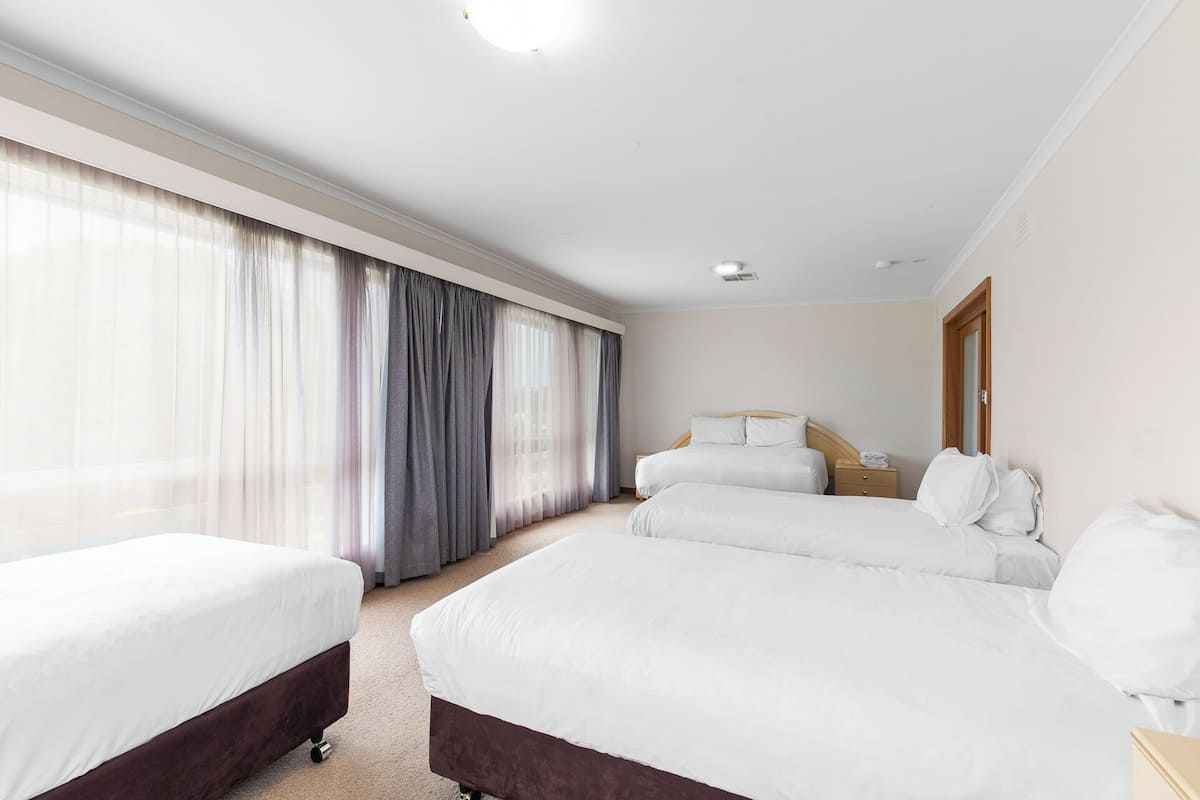 Une chambre d’hôtel avec deux lits simples et un lit double, une table de chevet en bois et une grande fenêtre avec des rideaux légers.