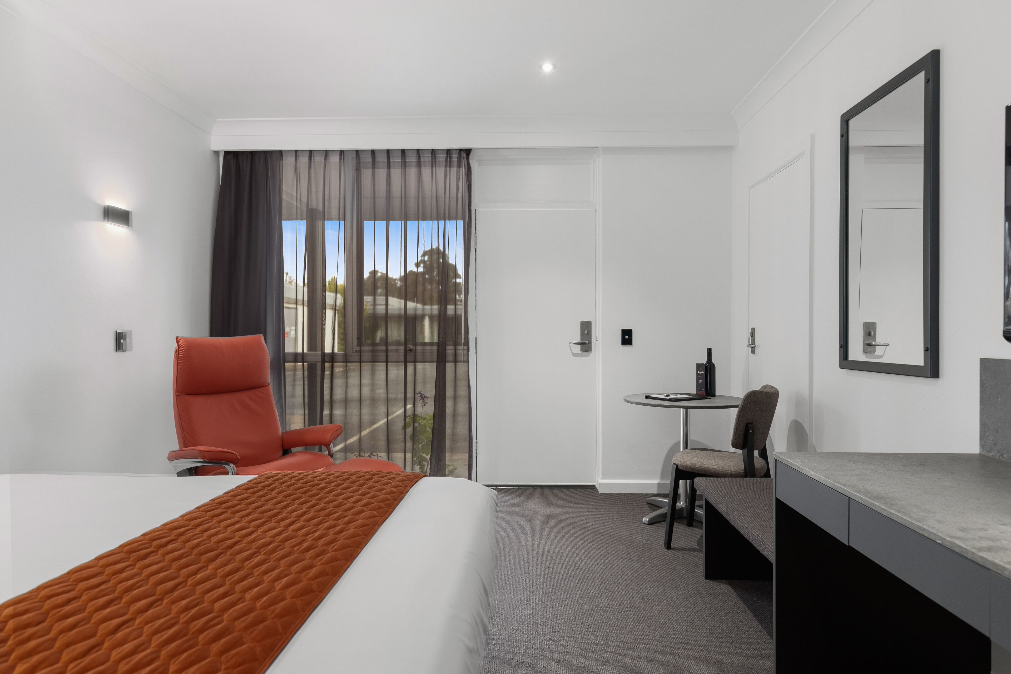 A hotel room with a bed, a desk, a chair, a small table, and a large mirror.