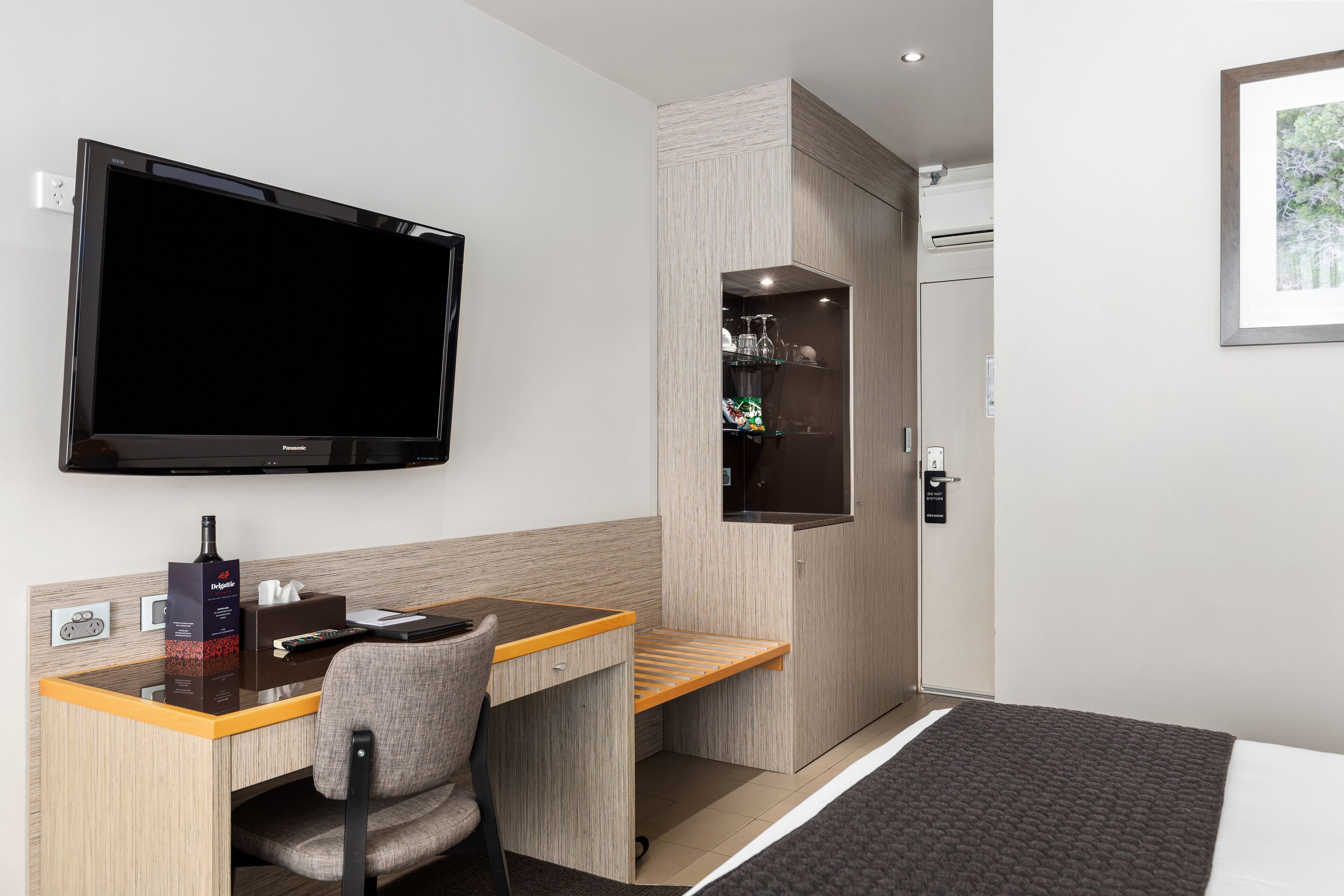 A hotel room with a wooden desk, a chair, a flat-screen TV, a minibar, and a bed with a dark cover.