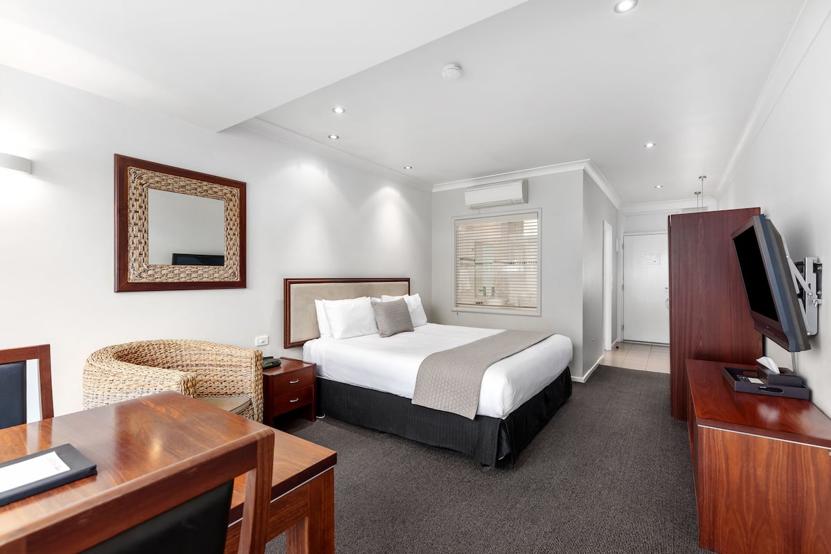 Une chambre d’hôtel équipée d’un lit, d’un bureau, d’une télévision, d’un miroir et d’une fenêtre avec des stores.