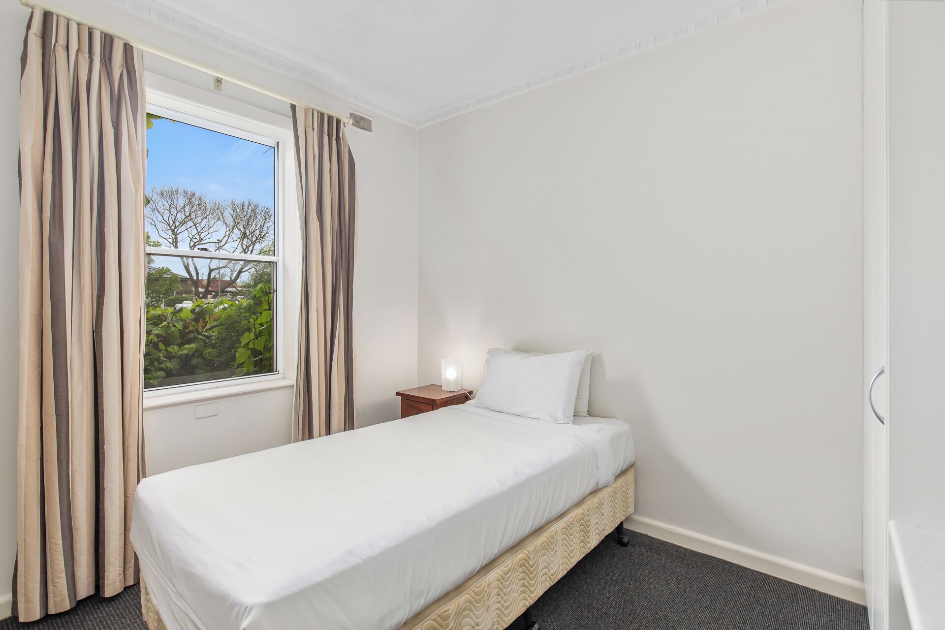 Una cama individual en una habitación con ventana, una mesita de noche con lámpara y vistas al exterior.