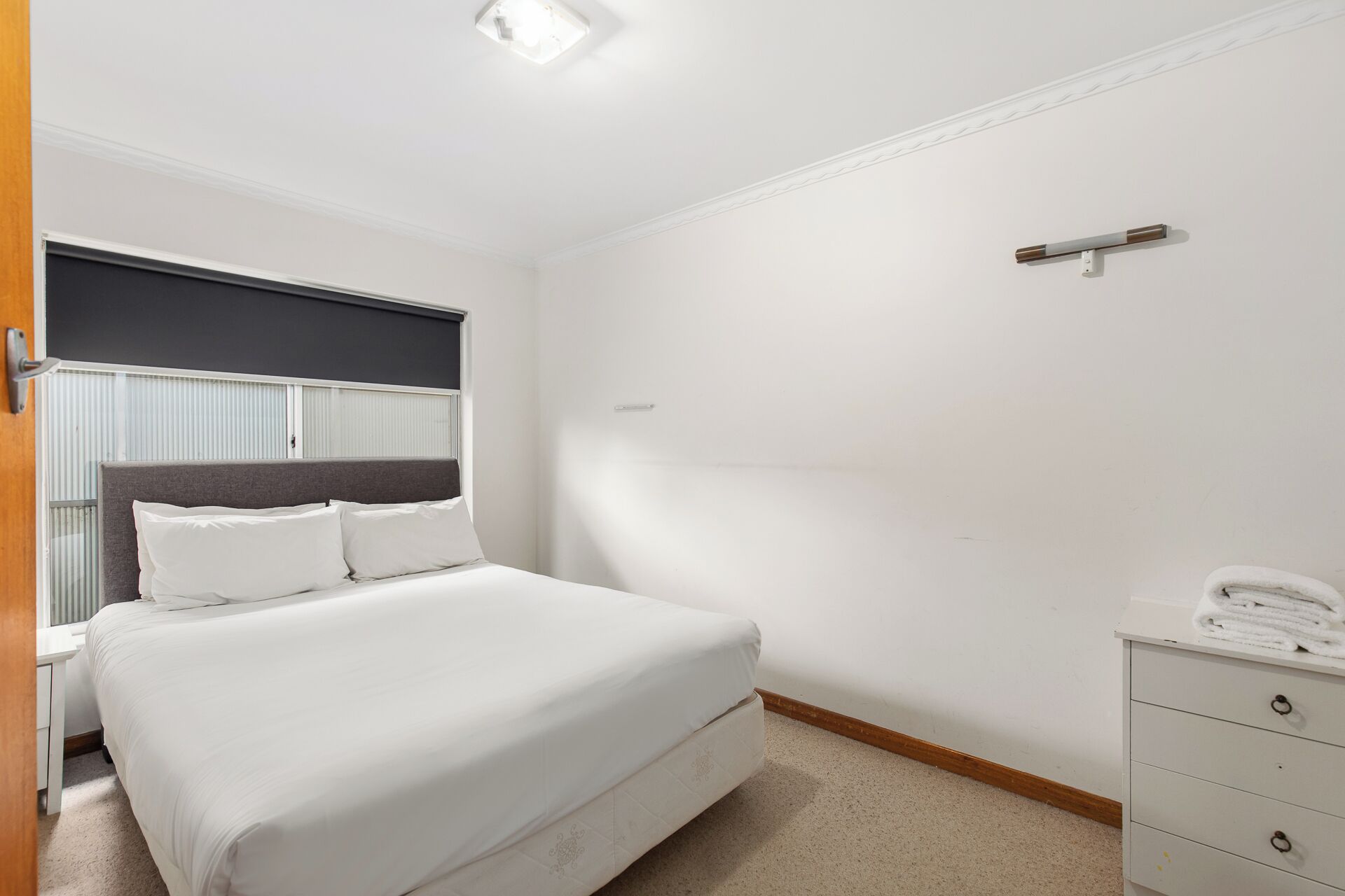 Een hotelkamer met een bed, nachtkastje, dressoir en een raam met jaloezieën.