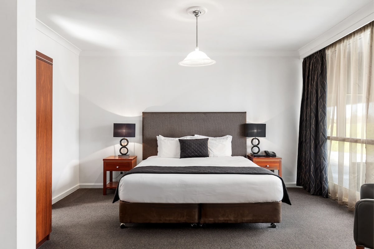 Une chambre d’hôtel avec un grand lit, deux tables de chevet avec des lampes, une armoire en bois et une fenêtre avec des rideaux légers.