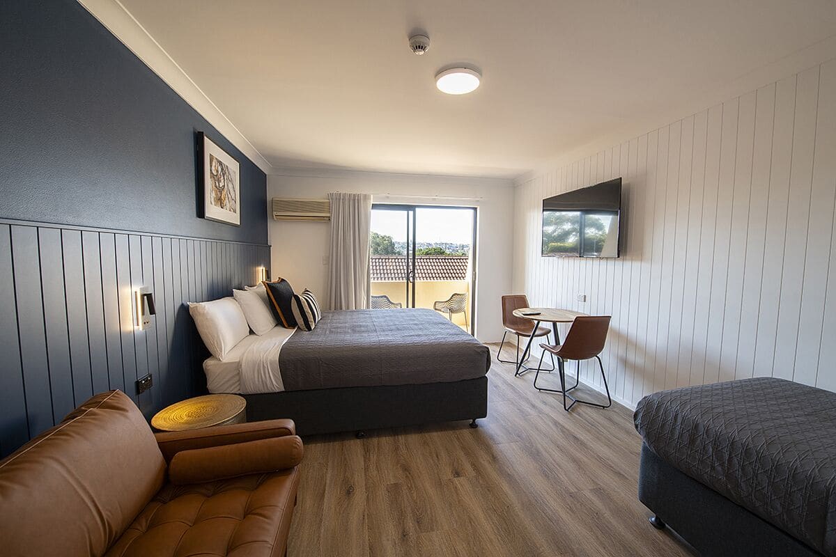A modern hotel room with a bed, a sofa, a small table, and a TV mounted on the wall.