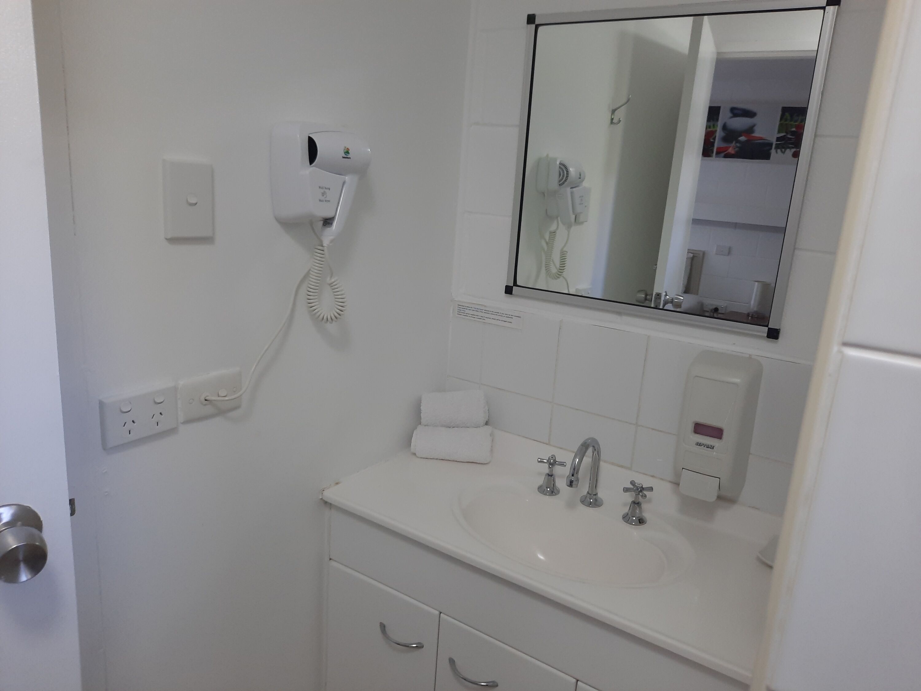 Un baño con lavamanos blanco, dos grifos, un espejo, un secador de pelo y una toalla.