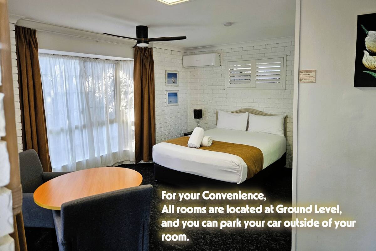 A hotel room with a bed, a table, and a chair. The room has a ceiling fan and a window with curtains.