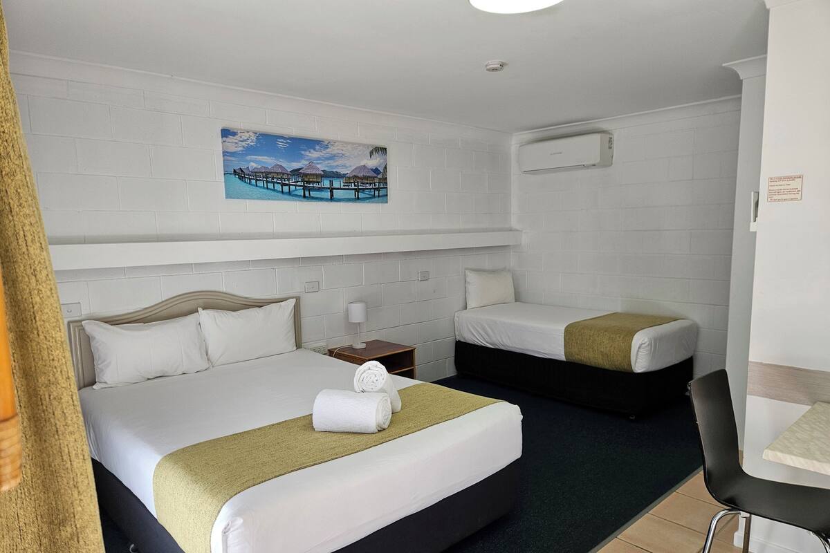 A hotel room with two beds, a painting of a beach scene, a white brick wall, and a small table with a chair.