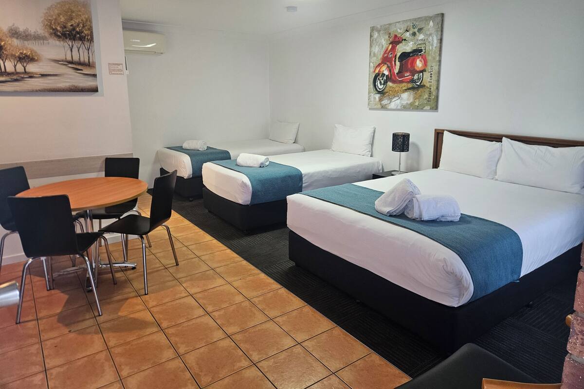 A hotel room with two beds, a dining table, and chairs. There is a painting on the wall and a lamp on the nightstand.