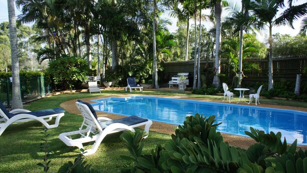 Piscina cercada por espreguiçadeiras brancas, área de churrasco e vegetação exuberante.