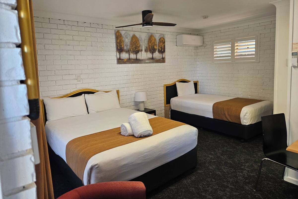 A hotel room with two beds, white brick walls, a ceiling fan, a painting, and a window with blinds.
