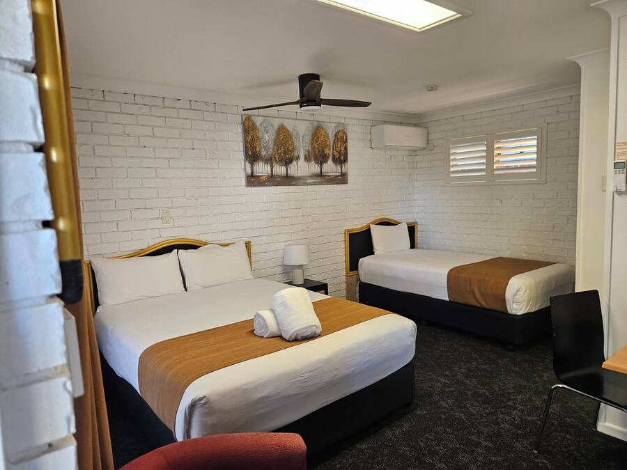 Une chambre d’hôtel avec deux lits, des murs en briques blanches, un ventilateur de plafond, un tableau et une fenêtre avec des stores.