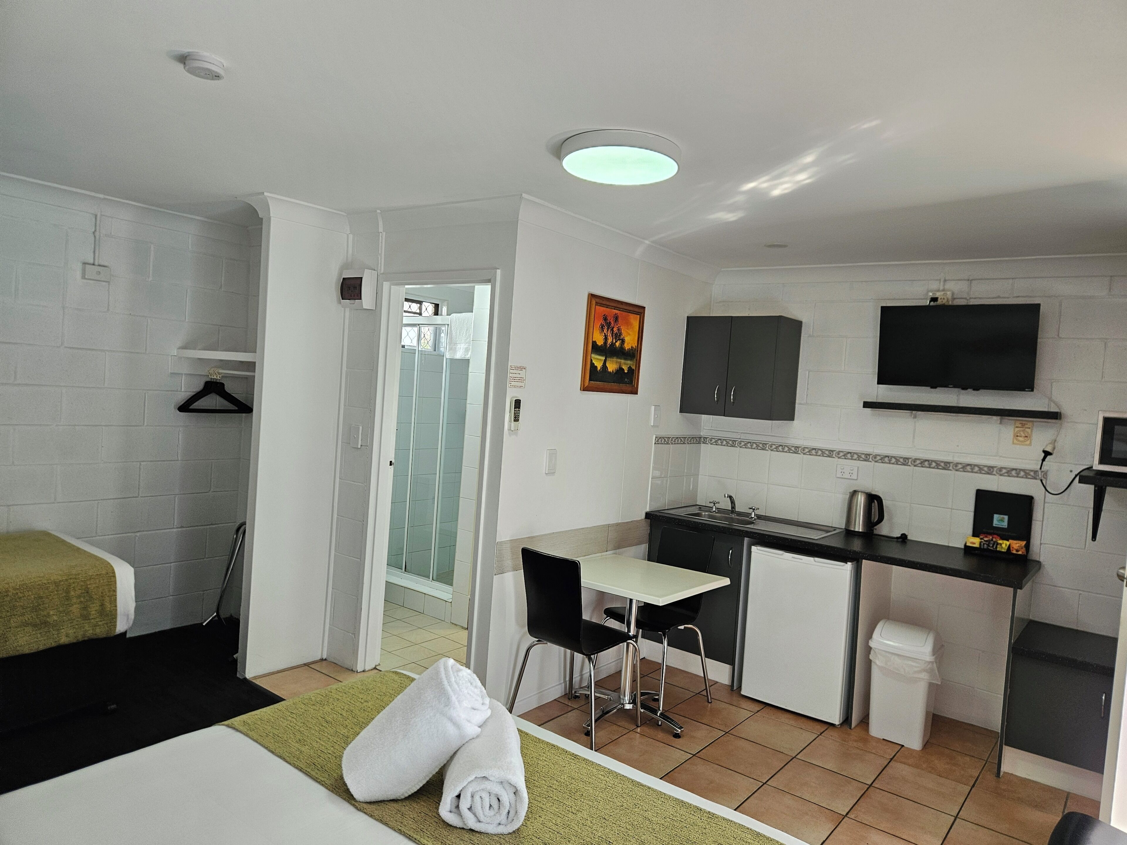 Habitación de hotel compacta con cama, escritorio, kitchenette pequeña y baño con ducha de vidrio.