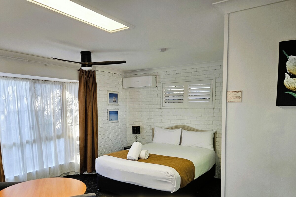 Ein Hotelzimmer mit einem Bett, einem Tisch und einem Stuhl. Das Zimmer verfügt über einen Deckenventilator und ein Fenster mit Vorhängen.