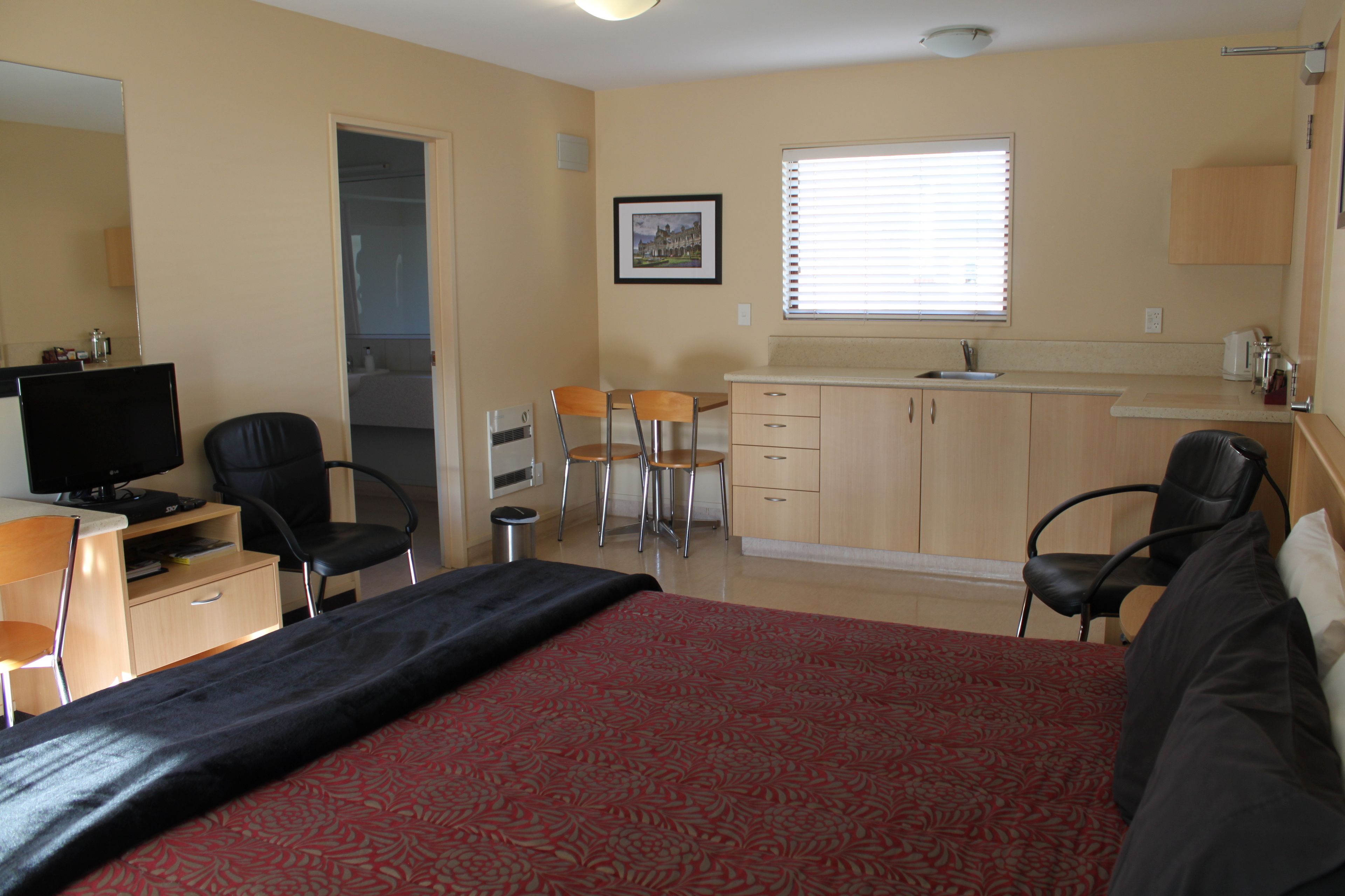 A hotel room with a bed, a desk with a television, two chairs, a small kitchenette, and a bathroom visible through an open door.