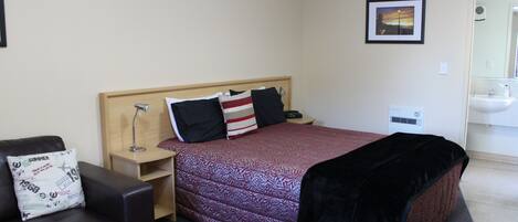 Habitación de hotel con cama, mesita de noche, lámpara, silla, almohada, manta, un cuadro en la pared y un baño con lavabo y espejo.