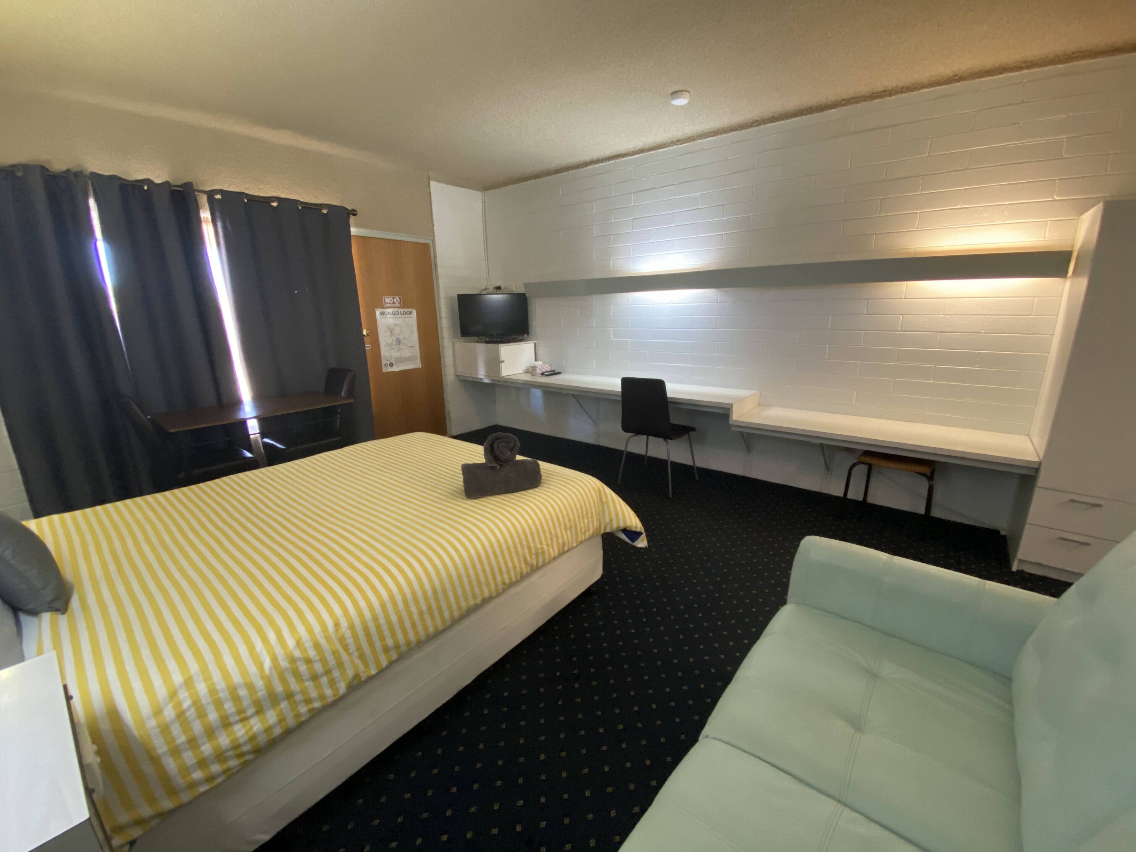 Quarto de hotel com cama, escrivaninha, cadeira, televisão e janela com cortinas.